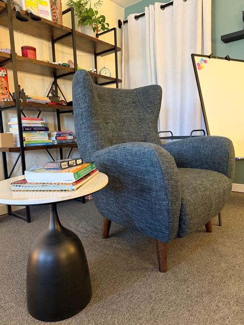Cozy consultation corner with a blue-gray wingback armchair, a small round side table with books, a tall black-framed shelf on the left, white curtains and a rolling whiteboard on the right